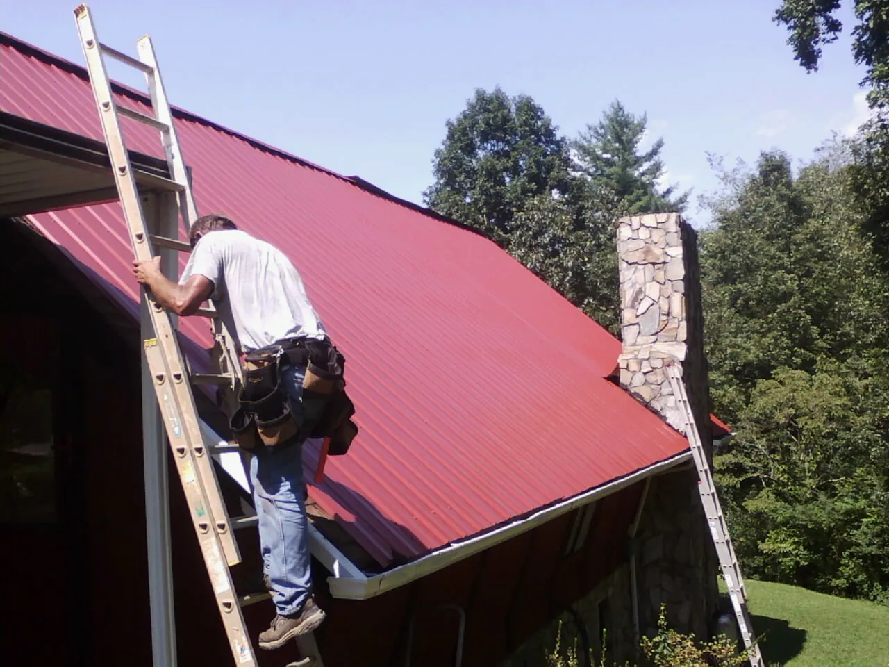 Roofer on a ladder beside a red metal roof and stone chimney — Soffit & Fascia Repair by Roofing Brothers in Traverse City
